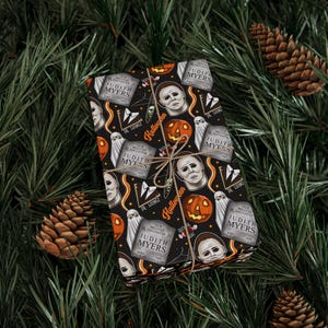 Puede incluir: Un regalo rectangular envuelto en papel negro con un patrón de Halloween que incluye ilustraciones de Michael Myers, calabazas naranjas, fantasmas blancos y lápidas con el nombre Judith Myers, atado con un cordel de color marrón.