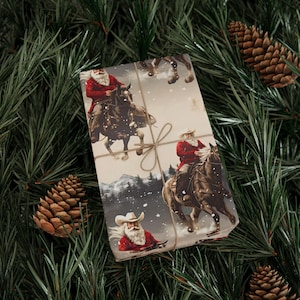 Puede incluir: Un regalo rectangular envuelto en papel con ilustraciones de Santa Claus con sombrero de vaquero y abrigo rojo montando a caballo en un paisaje nevado. El paquete está atado con un cordel marrón fino y presenta una escena invernal repetida.