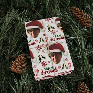 May include: A rectangular gift box wrapped in white paper featuring a repeating pattern of a person wearing a Santa hat, green Christmas trees, red snowflakes, and candy canes, with the text Team Jeremiah printed in red and green ink.