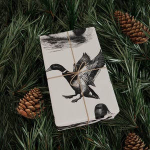 Pode incluir: Uma caixa de presente retangular embrulhada em papel branco sujo com uma ilustração a tinta preta de patos a voar e a nadar. O pacote está atado com um fio fino de cor castanha natural, criando um padrão de cruz simples.