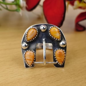 Könnte beinhalten: Ein silberner Ring in Form einer Hufeisenschnalle mit vier tropfenförmigen orangefarbenen Steinen in gezackten Fassungen. Die Metalloberfläche hat ein dunkel oxidiertes Finish mit drei kleinen polierten Silberkugeln zwischen den Steinen.