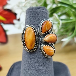 May include: A silver ring featuring a large oval orange stone set in a twisted rope border, positioned next to three smaller teardrop-shaped orange stones arranged vertically in a silver setting with a serrated edge detail.