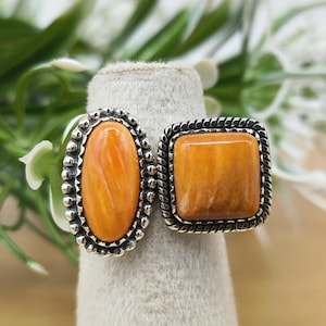 Könnte beinhalten: Ein Silberring mit zwei verschiedenen orangefarbenen Steinen in Metallfassungen. Ein Stein ist oval mit einem Perlenrand, der andere ist quadratisch mit einem gedrehten Kordelrand. Die Steine weisen natürliche Farbvariationen auf.