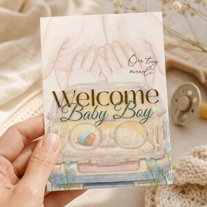 Könnte beinhalten: Eine rechteckige Grußkarte mit einer Aquarellillustration eines Neugeborenen im Krankenhaus-Inkubator, umrahmt von erwachsenen Händen. Der Text lautet "Our tiny miracle" und "Welcome Baby Boy" in goldener und dunkelblauer Schreibschrift.