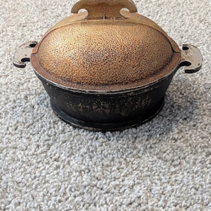 May include: A round, black metal pot with a textured, gold-colored domed lid. The lid features a central handle, and the pot has two side handles. The exterior of the pot and lid shows a weathered, speckled finish with visible signs of wear.
