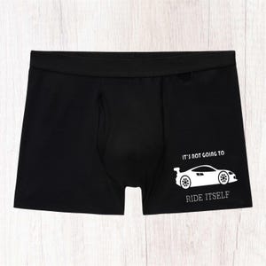 May include: Black boxer briefs featuring a white graphic of a sports car on the right leg. Above the car, the text reads IT'S NOT GOING TO, and below the car, the text reads RIDE ITSELF. The garment has a wide elastic waistband and a front pouch.