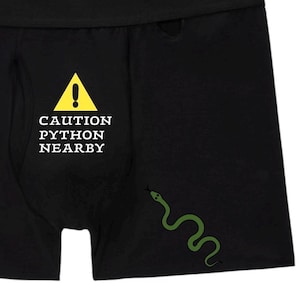 Peut inclure: Cale&ccedil;on noir avec un panneau d'avertissement triangulaire jaune portant un point d'exclamation et le texte CAUTION PYTHON NEARBY en lettres capitales blanches. Une petite illustration de serpent vert est situ&eacute;e sous le texte.