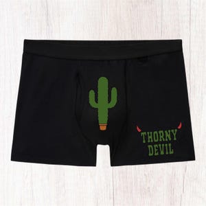 May include: Black boxer briefs featuring a central green cactus graphic in a brown pot. The right side displays the text "THORNY DEVIL" in green block letters, accented by two small red devil horns positioned above the word "THORNY."