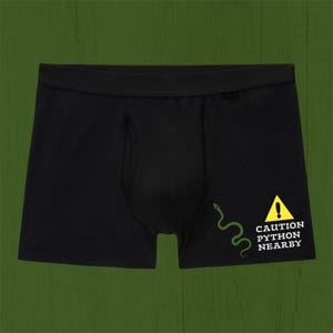 May include: Black boxer briefs featuring a graphic of a green snake and a yellow triangular warning sign with an exclamation point. Below the sign, the text reads CAUTION PYTHON NEARBY in white capital letters on the right side.