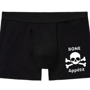 May include: Black boxer briefs featuring a white graphic print of a skull and crossbones on the right leg. Above the skull is the word BONE and below the skull is the word Appétit in white text, creating the phrase BONE Appétit on the fabric.