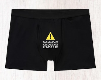 Peut inclure: Un caleçon noir avec un triangle jaune d'avertissement contenant un point d'exclamation et le texte blanc CAUTION CHOKING HAZARD! imprimé sur le panneau central avant. Le vêtement possède une large ceinture élastique et une coupe standard.