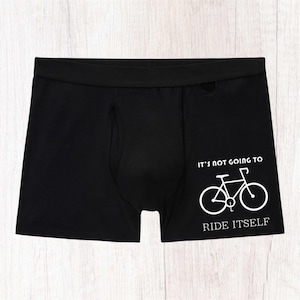 May include: Black boxer briefs featuring a white graphic of a bicycle on the right side. Above the bicycle, the text reads IT'S NOT GOING TO, and below the bicycle, the text reads RIDE ITSELF. The garment has a wide elastic waistband.