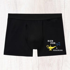 May include: Black boxer briefs featuring a yellow graphic of a magic lamp with blue smoke swirls. The text printed in white reads RUB FOR A SURPRISE. The garment has a wide elastic waistband and a contoured front pouch design for a fitted look.