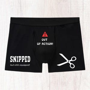 May include: Black boxer briefs featuring a red warning triangle with an exclamation point, the white text OUT OF ACTION!, SNIPPED, but still equipped!, and a white graphic of open scissors on the lower right side of the garment.