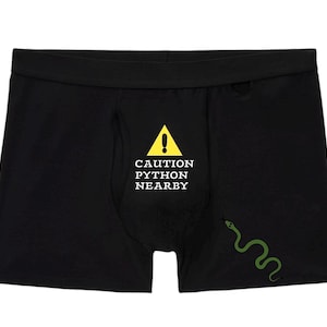 Peut inclure: Cale&ccedil;on noir arborant un panneau de signalisation triangulaire jaune avec un point d'exclamation au-dessus du texte blanc CAUTION PYTHON NEARBY. Une petite illustration de serpent vert est situ&eacute;e sur le bas droit du tissu du v&ecirc;tement.