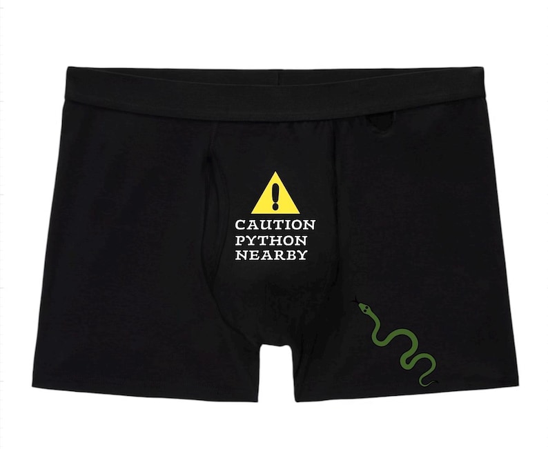 Peut inclure: Cale&ccedil;on noir arborant un panneau de signalisation triangulaire jaune avec un point d'exclamation au-dessus du texte blanc CAUTION PYTHON NEARBY. Une petite illustration de serpent vert est situ&eacute;e sur le bas droit du tissu du v&ecirc;tement.