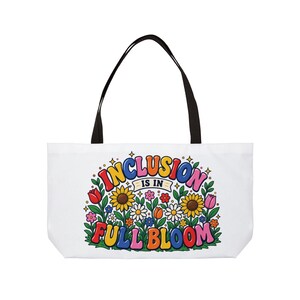 May include: A white tote bag with black handles featuring a colorful graphic of various flowers and the text "INCLUSION IS IN FULL BLOOM" in a bold, multi-colored font. The design includes sunflowers, tulips, and daisies with green leaves.