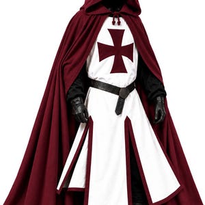 Puede incluir: Una capa con capucha de color burdeos intenso sobre una túnica blanca con un emblema de cruz roja oscura en el pecho. El conjunto incluye un cinturón negro con hebilla circular, prendas negras de manga larga, guantes y botas de cuero marrón.
