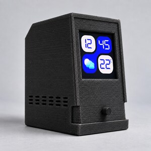 May include: A black 3D-printed digital desktop clock with a textured finish. The front features a square screen displaying the time 12:45, a weather icon showing clouds, and a temperature of 22 degrees Celsius. A small black circular knob is below.