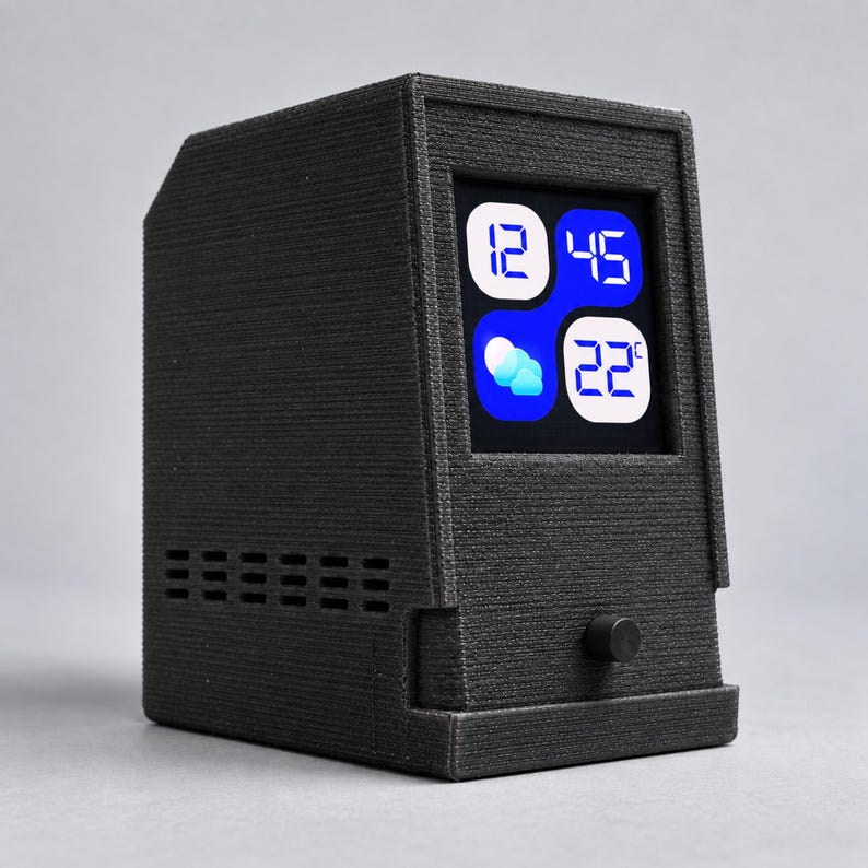 May include: A black 3D-printed digital desktop clock with a textured finish. The front features a square screen displaying the time 12:45, a weather icon showing clouds, and a temperature of 22 degrees Celsius. A small black circular knob is below.