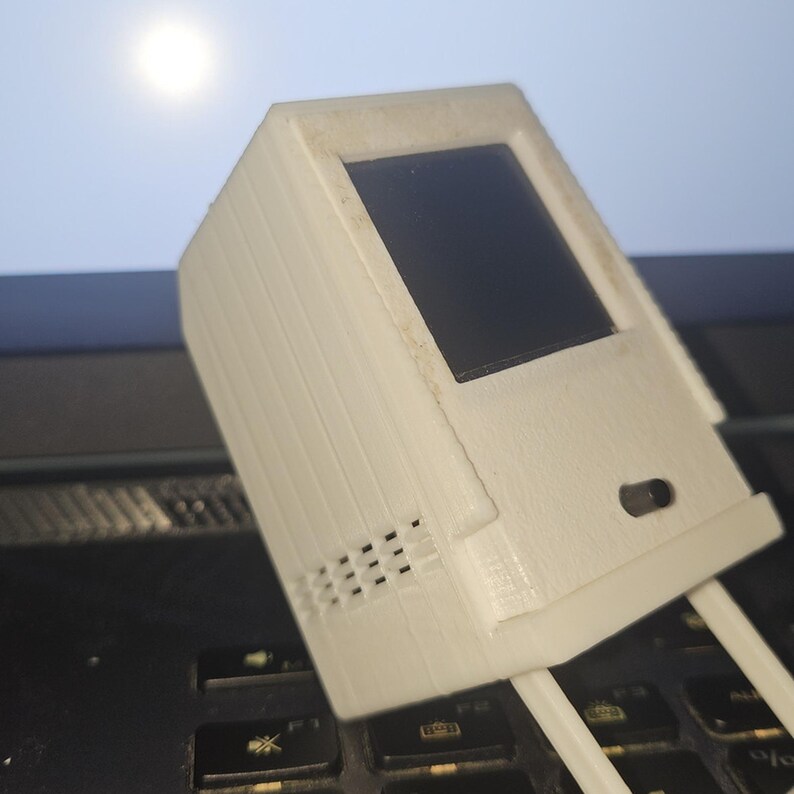 May include: A white, 3D-printed miniature computer model featuring a rectangular screen area, a small circular button on the front panel, and a series of horizontal ventilation slits along the side, supported by two thin, parallel white legs.