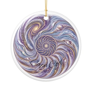 May include: A round white ceramic ornament featuring a swirling purple, blue, and gold cosmic design with a central nautilus shell pattern, scattered small seashells, and the text Cape Cod written in a black cursive script with a gold hanging cord.