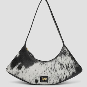 以下が含まれることがあります： 黒と白の牛革模様が特徴の三日月型ショルダーバッグ。黒いストラップが1本、ゴールドのジッパー、バッグ下部中央には「Luna」というゴールドの文字がプリントされた小さな黒い長方形のパッチが付いています。