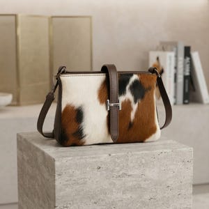 以下が含まれることがあります： 白、茶、黒の3色のカウハイド柄が特徴の長方形のショルダーバッグ。ダークブラウンのレザーストラップが、H型のシルバーメタルのバックルでフロントフラップを固定しており、両サイドにも同色のストラップが付いています。