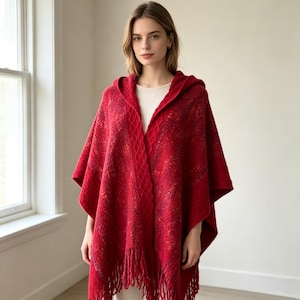 Op de afbeelding: Een felrode poncho met capuchon, voorzien van een gestructureerd breipatroon met kabelbreidetails langs de voorranden en een zoom met franjes. Het kledingstuk wordt gedragen over een effen lichtgekleurde top en heeft een dikke, warme stof.