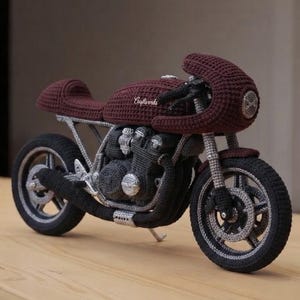Puede incluir: Un modelo de motocicleta tejido a ganchillo en hilo color burdeos oscuro y gris carbón. La pieza presenta un carenado frontal redondeado, un tanque de combustible con el texto bordado Craftworks, un bloque de motor texturizado, neumáticos negros y llantas plateadas.