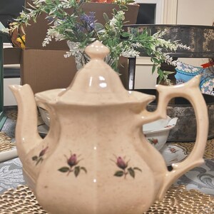 May include: A cream-coloured ceramic teapot with a speckled finish and a pointed lid. The teapot features a floral pattern of small pink rosebuds with green leaves on the body, a curved handle, and a spout, all resting on a hexagonal base.