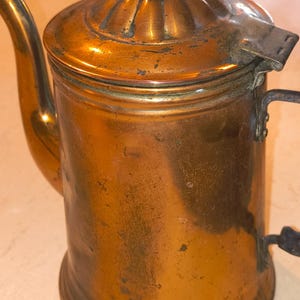 May include: A vintage copper coffee pot featuring a cylindrical body, a curved spout, and a hinged lid topped with a decorative wooden finial. The metal surface displays a warm, weathered patina with visible signs of age, oxidation, and surface wear.