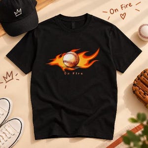 May include: A black short-sleeve t-shirt featuring a graphic of a baseball surrounded by orange and yellow flames. Below the baseball, the text "On Fire" is printed in a small, simple font. The shirt has a crew neckline and a standard fit.