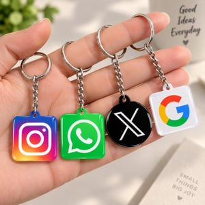 Może przedstawiać: Cztery metalowe breloki z logo mediów społecznościowych i technologii. Pierwszy to kwadratowa ikona Instagrama, drugi to kwadratowa ikona WhatsAppa, trzeci to okrągłe czarne logo X, a czwarty to kwadratowe logo Google G, wszystkie ze srebrnymi kółkami.