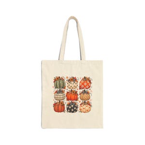 Peut inclure: Un sac fourre-tout en toile beige avec une impression graphique centrale représentant neuf citrouilles stylisées disposées en grille. Chaque citrouille présente des motifs uniques, notamment des rayures, des pois et des carreaux, ornés de tournesols et de feuilles d'automne.