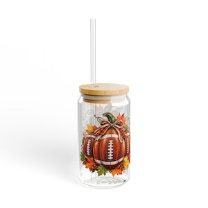 May include: A clear glass tumbler with a bamboo lid and a clear straw. The glass features a graphic of a football shaped like a pumpkin, accented with a plaid bow, surrounded by colorful autumn leaves in shades of orange, red, and yellow.