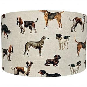 Può includere: Un paralume cilindrico caratterizzato da un motivo ripetuto di varie razze canine stampate su un tessuto testurizzato color beige chiaro. I cani sono raffigurati in molteplici pose e colori, tra cui marrone, nero, bianco e marrone chiaro.