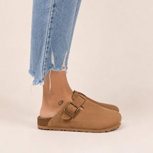 May include: A tan suede slip-on clog featuring a rounded toe, a cork-textured midsole, and an adjustable strap across the top secured with a bronze-toned metal buckle. The footwear has a flat sole and a simple, functional design for casual wear.