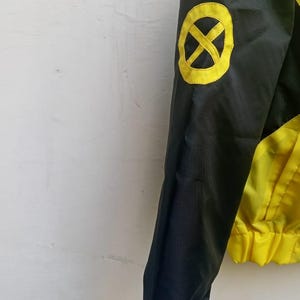 Może przedstawiać: Czarno-ż&oacute;łta kurtka z długim rękawem, na kt&oacute;rym znajduje się okrągły ż&oacute;łty emblemat z czarnym symbolem X na ramieniu. Kurtka posiada czarny rękaw oraz ż&oacute;łtą dolną część z elastycznym ściągaczem w pasie.