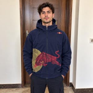 Könnte beinhalten: Eine marineblaue Kapuzenjacke mit Reißverschluss, die auf der Vorderseite eine große rote Stiergrafik, ein goldenes Kreismuster hinter dem Stier und ein kleines Red Bull-Logo auf der linken Brust zeigt. Die Jacke hat lange Ärmel und einen Reißverschluss.