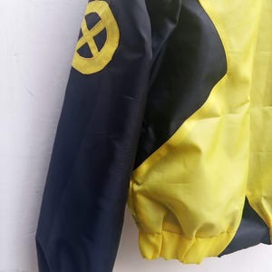 Może przedstawiać: Ż&oacute;łto-czarna kurtka z okrągłym ż&oacute;łtym emblematem z czarnym krzyżem na g&oacute;rnej części rękawa. Odzież posiada długie rękawy z prążkowanymi mankietami, jednolity czarny panel rękawa oraz jaskrawoż&oacute;łtą część gł&oacute;wną korpusu.