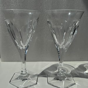 May include: Two clear crystal wine glasses featuring a flared bowl with vertical cut panels and a distinct hexagonal base. The stem is faceted, leading down to a flat, six-sided foot, providing a stable foundation for each glass.