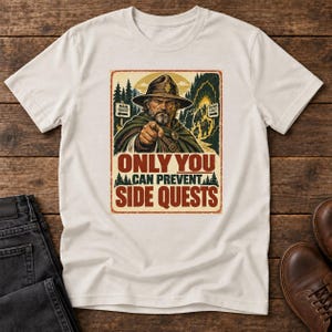 Könnte beinhalten: Ein cremefarbenes T-Shirt mit einer Grafik eines bärtigen Abenteurers mit Hut, der nach vorne zeigt. Das Design zeigt einen Waldhintergrund, Schilder mit der Aufschrift Main Quest und Shiny Loot sowie den Text Only You Can Prevent Side Quests.