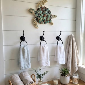May include: A wall-mounted sea turtle sculpture crafted from iridescent abalone shells, positioned above three black metal towel hooks. Each hook holds a white towel, while a beige waffle-weave towel hangs to the right. A wicker basket holds folded towels.