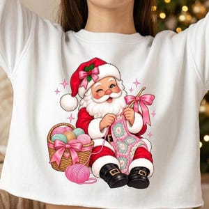May include: A white long-sleeve sweatshirt featuring a graphic of Santa Claus winking while crocheting a pink and green granny square blanket. Santa wears a red suit with white trim and black boots, sitting next to a wicker basket filled with yarn.