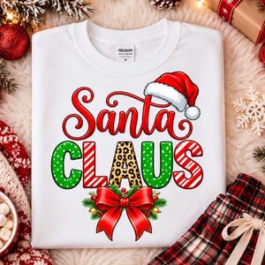 May include: A white t-shirt featuring the text Santa CLAUS in red, green, and leopard print. The word Santa is topped with a red and white Santa hat. The word CLAUS is accented with a red bow with green pine needles and red berries.