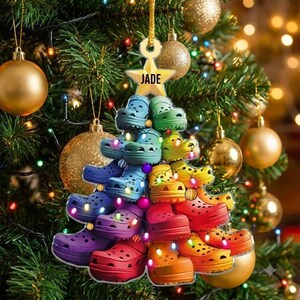 May include: A Christmas tree ornament shaped like a pyramid of colorful clogs in a rainbow gradient. The ornament features a gold star at the top with the name JADE printed in black text. Small multicolored lights are interspersed among the clogs.