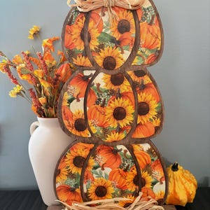 May include: A vertical wooden decoration featuring three stacked pumpkin shapes with a dark brown frame. The surface displays a vibrant pattern of orange pumpkins and yellow sunflowers. A raffia bow sits at the top, and raffia rests at the base.
