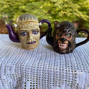 May include: Two ceramic teapots shaped like monster heads. The left teapot features a pale, stitched face with a purple handle and spout. The right teapot depicts a brown, furry werewolf face with a black handle and spout, showing sharp teeth.