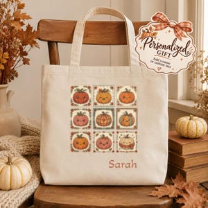 Peut inclure: Un sac fourre-tout en toile couleur crème orné d'une grille de neuf citrouilles illustrées aux expressions et motifs variés. Le prénom Sarah est imprimé en orange sous la grille. Une étiquette décorative avec un nœud indique Personalized GIFT.